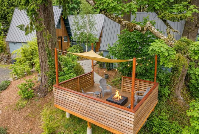 Elevated wooden deck with Adirondack chairs and a glowing fire pit under a tan shade sail and string lights, nestled among lush green trees in front of modern A-frame cabins with metal roofs