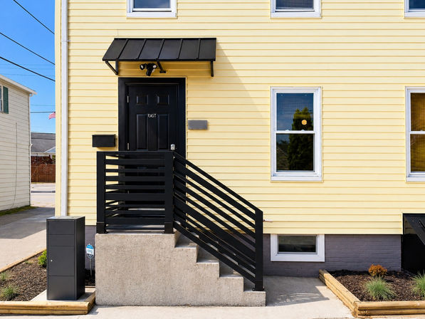 Sunny yellow clapboard house front showing a black door under a metal awning, concrete steps with modern black slatted railing, white-trim windows, mailbox and security camera.