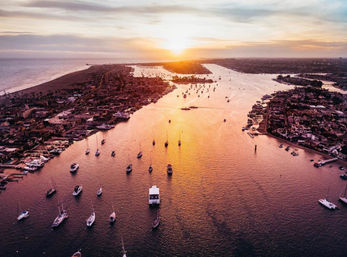 Aerial sunset over a coastal channel, golden light reflecting on a harbor filled with anchored sailboats, yachts and waterfront neighborhoods.