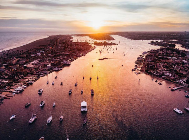 Aerial sunset over a coastal channel, golden light reflecting on a harbor filled with anchored sailboats, yachts and waterfront neighborhoods.