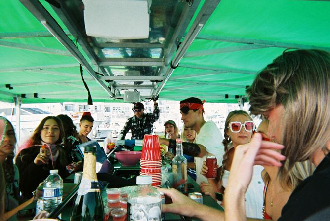 Group of friends enjoying a lively boat party under a green canopy at a marina, gathered around a bar-style table with red solo cups, bottles and sunglasses