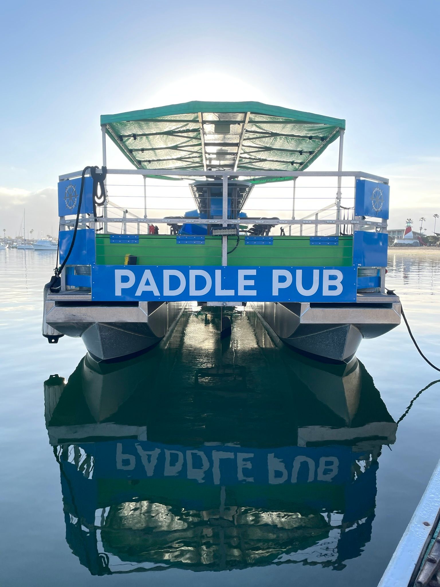Blue-green paddle pub party boat docked at a calm marina at sunrise, sunlit canopy and crisp reflection on the glassy harbor water.