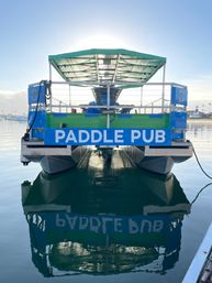 Blue-green paddle pub party boat docked at a calm marina at sunrise, sunlit canopy and crisp reflection on the glassy harbor water.