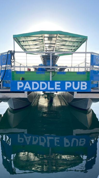 Blue-green paddle pub party boat docked at a calm marina at sunrise, sunlit canopy and crisp reflection on the glassy harbor water.