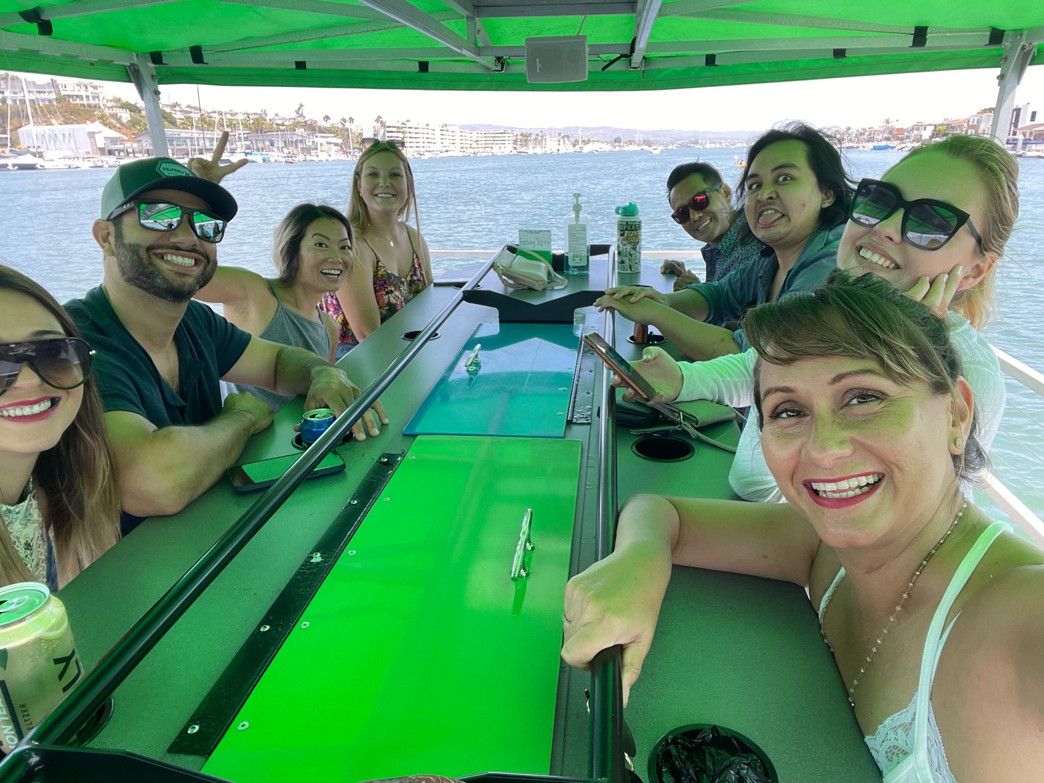 Smiling group of friends on a green-canopied party boat gathered around a central bar with drinks and sunglasses, harbor marina and waterfront homes visible in the background.