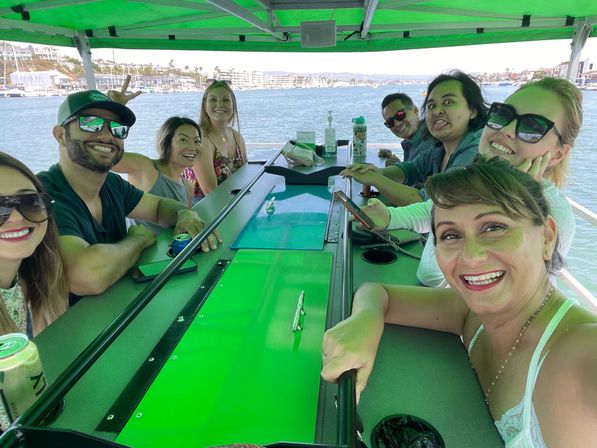 Smiling group of friends on a green-canopied party boat gathered around a central bar with drinks and sunglasses, harbor marina and waterfront homes visible in the background.