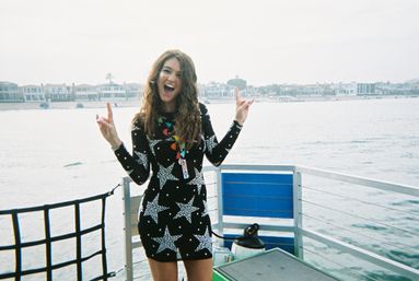 Smiling woman in a black star-patterned dress striking a celebratory pose on a boat deck by the waterfront, with coastal homes and calm water in the background.
