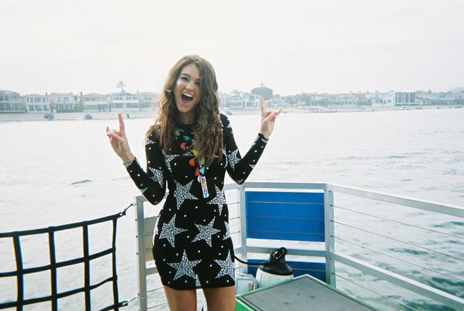 Smiling woman in a black star-patterned dress striking a celebratory pose on a boat deck by the waterfront, with coastal homes and calm water in the background.