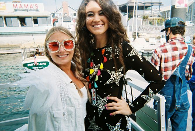Two smiling women in festive outfits on a sunny waterfront pier; one wears oversized sparkly sunglasses, the other a star-patterned dress with colorful tassels, boats and marina buildings in the background.