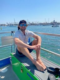 Relaxed man in sunglasses and backward cap sitting barefoot on a boat bench at a sunny coastal marina, with sailboats, palm trees and waterfront homes in the background.
