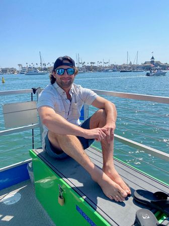 Relaxed man in sunglasses and backward cap sitting barefoot on a boat bench at a sunny coastal marina, with sailboats, palm trees and waterfront homes in the background.