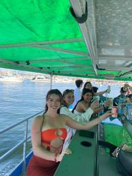 Group of friends cheering with canned drinks at a covered bar on a party boat in a sunny marina with yachts and calm water in the background