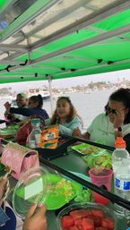 Group of friends sharing snacks under a bright green canopy on a pontoon boat in a marina, plates of watermelon, cheese and chips with waterfront homes and yachts in the background