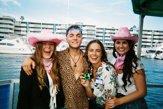 Four friends laughing on a boat at a sunny marina — two women in pink cowboy hats and bandanas, a man in a leopard-print shirt with sunglasses, and a woman holding a green disposable camera with yachts and waterfront condos in the background.