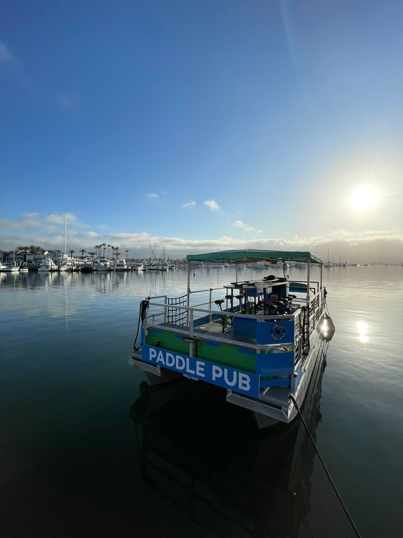 Sunrise over a calm marina with a paddle-pub style pontoon moored in the foreground, mirrored harbor water, sailboats and palm trees along the shoreline under a clear blue sky.
