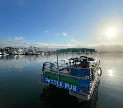 Sunrise over a calm marina with a paddle-pub style pontoon moored in the foreground, mirrored harbor water, sailboats and palm trees along the shoreline under a clear blue sky.