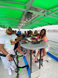 Smiling group enjoying drinks and snacks around a central bar on a green-canopied pedal party boat cruising a marina-lined harbor.