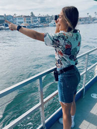 Smiling person on a boat deck wearing a floral shirt and denim shorts, pointing and holding binoculars toward a marina of boats and waterfront homes across a sunny harbor.