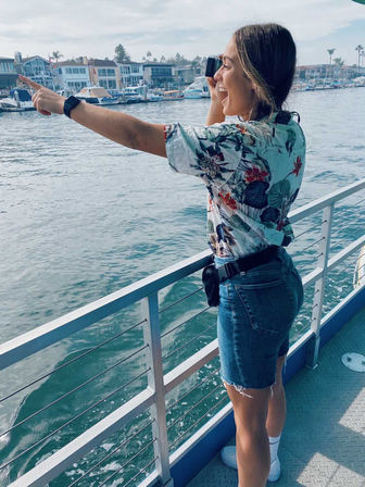 Smiling person on a boat deck wearing a floral shirt and denim shorts, pointing and holding binoculars toward a marina of boats and waterfront homes across a sunny harbor.
