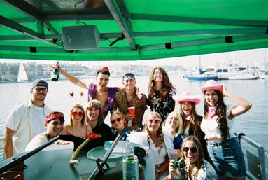 Group of friends at a sunny boat party under a green canopy in a marina, celebrating with drinks and pink cowboy hats, yachts and sailboats in the background.
