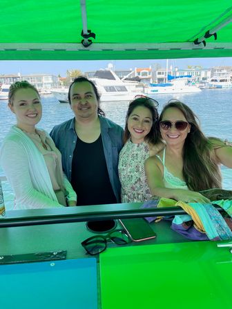 Four smiling friends on a covered boat with a bright green canopy, posing at a sunny marina with yachts and waterfront homes in the background — casual summer outing on the water.
