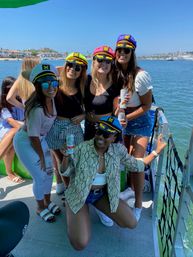 Five friends in colorful captain hats smiling on a sunny boat party in a coastal marina, holding canned drinks with yachts and waterfront homes in the background.