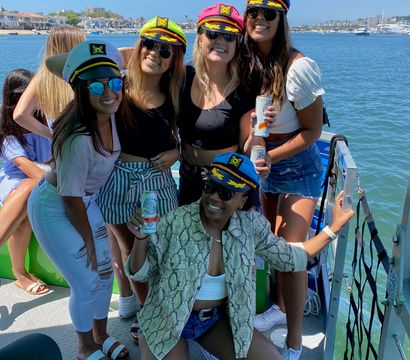 Five friends in colorful captain hats smiling on a sunny boat party in a coastal marina, holding canned drinks with yachts and waterfront homes in the background.