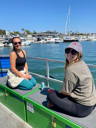 Two friends smiling on a sunny coastal marina dock, sitting on green storage benches by blue harbor water with yachts and sailboats moored under a clear sky.