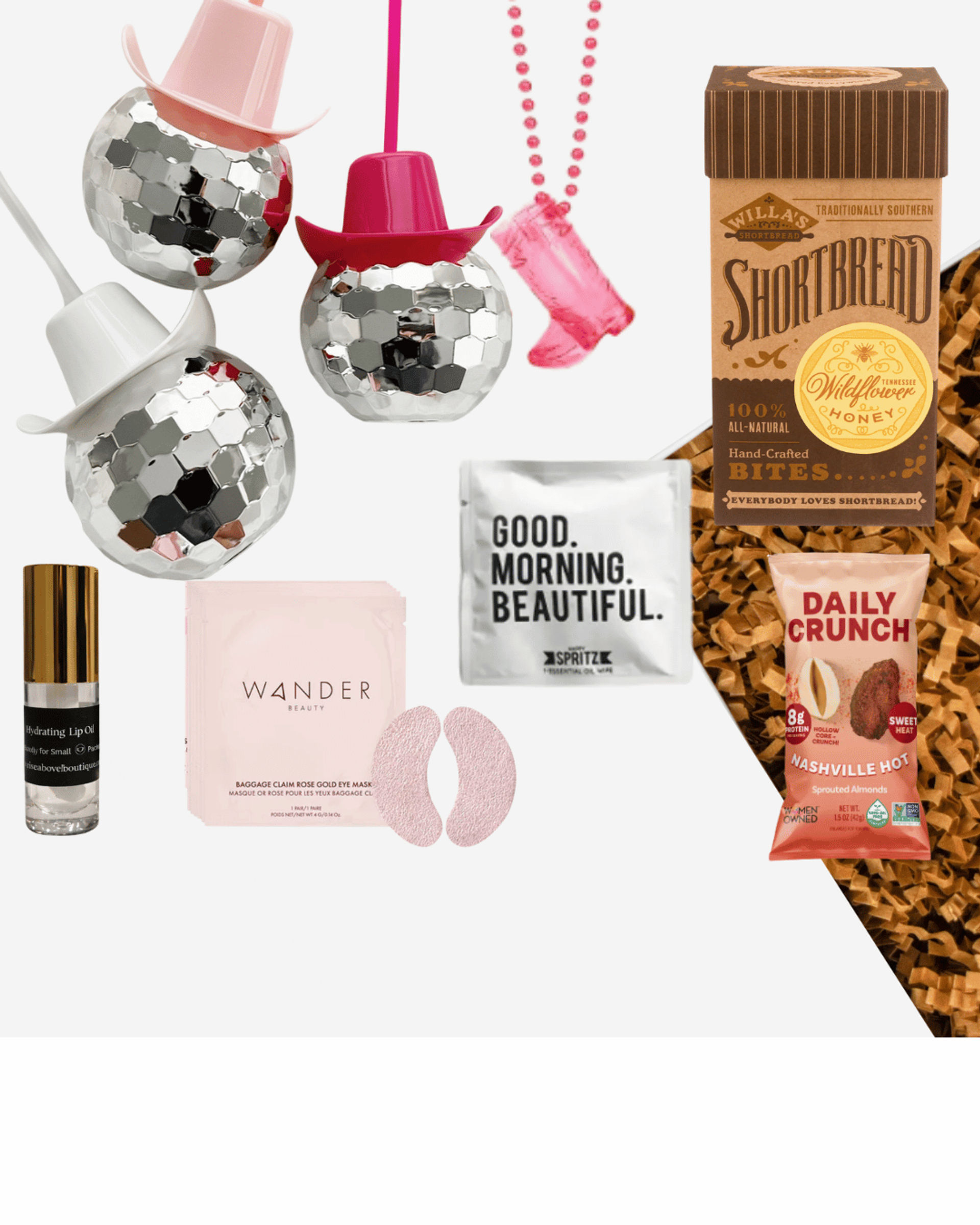 Flat-lay gift box on white background with mirrored disco-ball ornaments wearing tiny cowboy hats, pink beaded cowgirl necklace, shortbread cookie box, Nashville-style spicy almond snack, hydrating lip oil, rose under-eye gel patches with pink packet, and a sachet reading ‘Good. Morning. Beautiful.’ over brown crinkle filler.