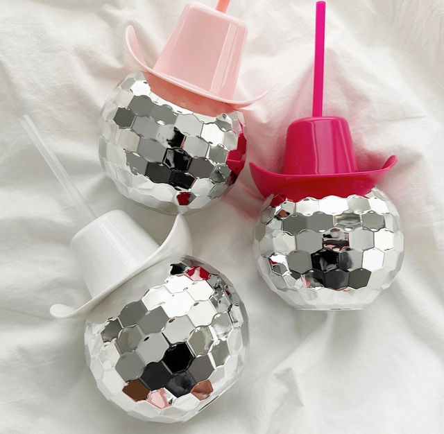 Four mirrored disco-ball tumblers with hexagonal facets, each topped with a plastic cowboy-hat lid and straw in pink, hot pink, pale pink and white, arranged on a white sheet — playful party drinkware.