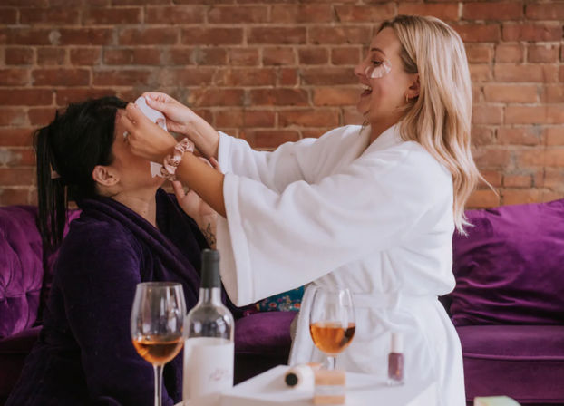 Two friends in bathrobes enjoying an at-home spa party, one applying under-eye patches to the other while they laugh, with wine glasses, a bottle and skincare items on a table and a purple couch against an exposed brick wall.