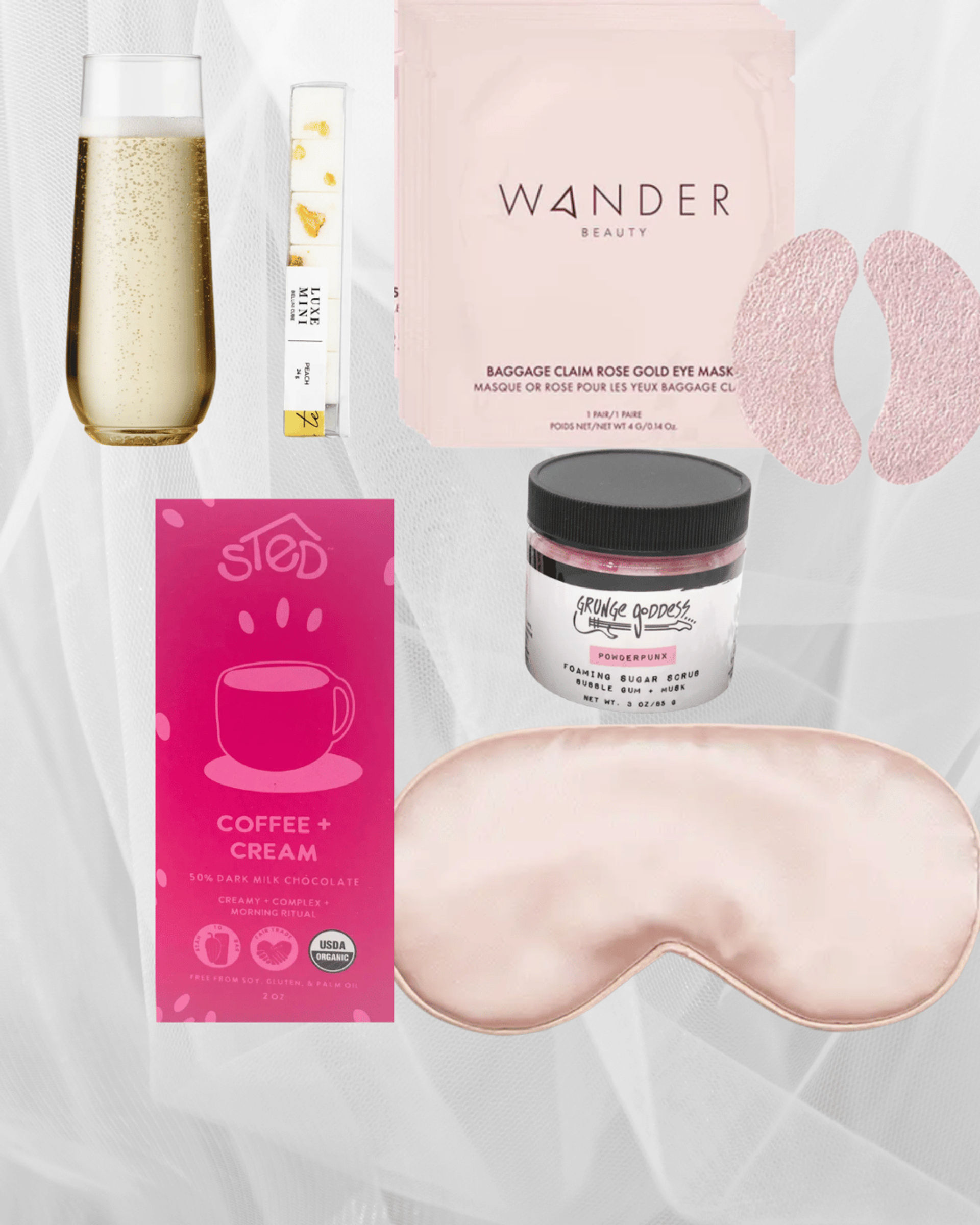 Pink-themed self-care flatlay with champagne flute, gold-flecked serum vial, rose-gold eye patches, silk sleep mask, coffee-and-cream chocolate bar, and jar of foaming sugar scrub on soft white fabric.