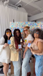 Four smiling women in sunglasses pose indoors at a holographic sequin photo-booth backdrop, holding colorful instant photos and a cupcake with a lit candle.