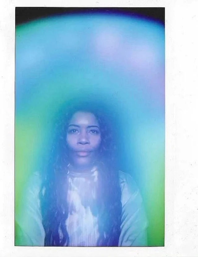 Portrait of a person with long dark hair and a calm expression, surrounded by a vivid blue-green-purple aura and soft ethereal glow in a Polaroid-style frame.
