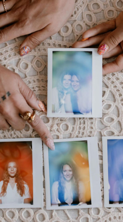 Flat lay of hands with rings and nail art arranging five instant Polaroid portraits on a lace tablecloth, featuring soft-focus smiling portraits with colorful light leaks and a playful vintage vibe.
