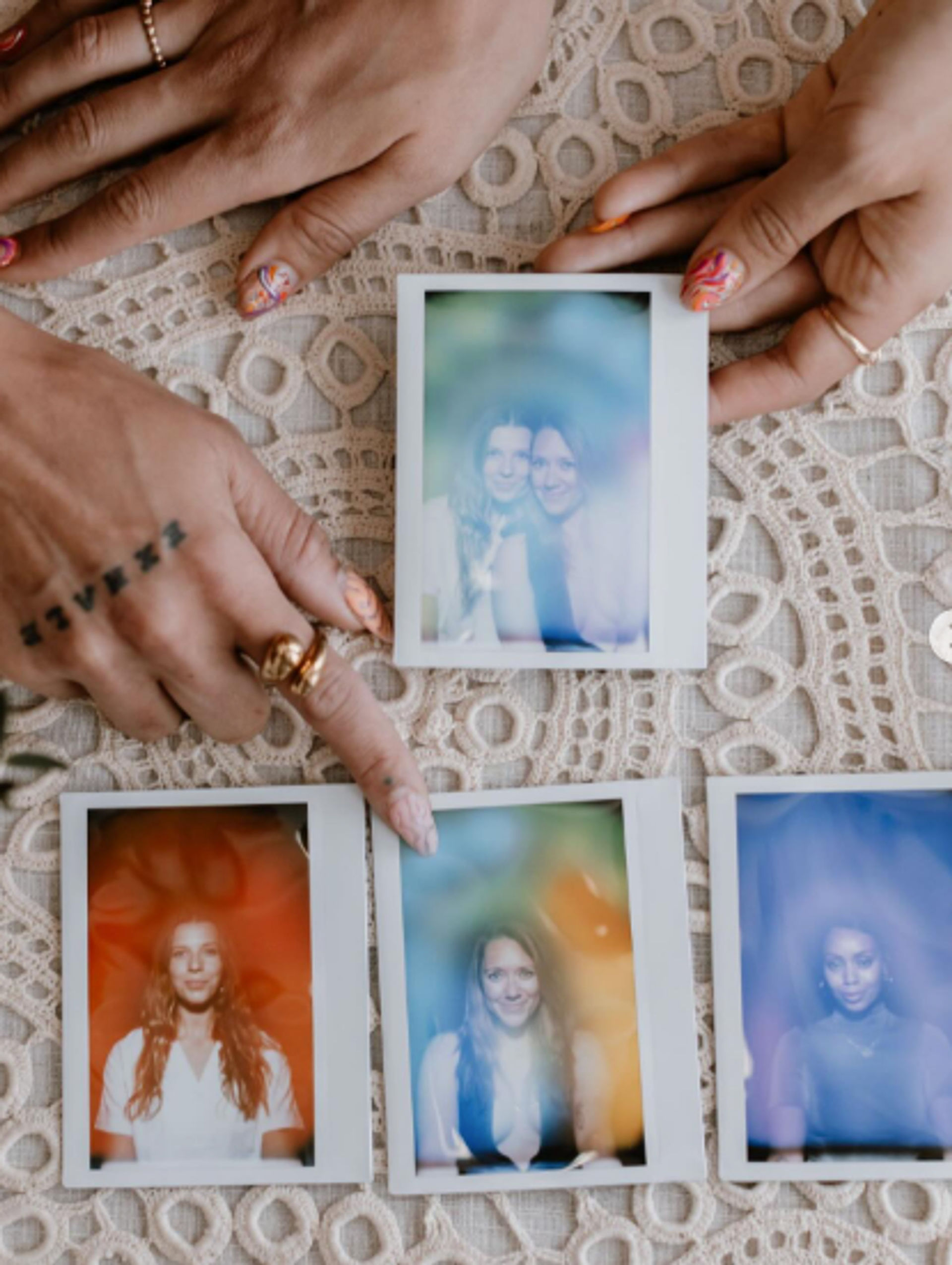 Flat lay of hands with rings and nail art arranging five instant Polaroid portraits on a lace tablecloth, featuring soft-focus smiling portraits with colorful light leaks and a playful vintage vibe.