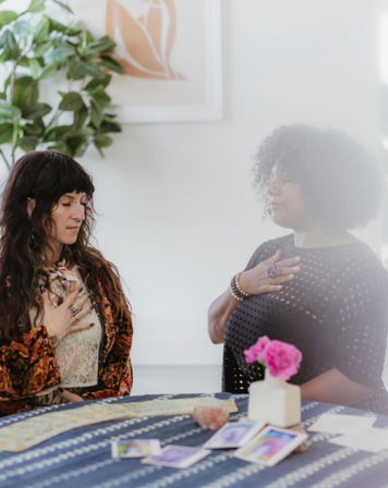 Two women in a cozy indoor spiritual session, hands over their hearts during a calm tarot reading with tarot cards, crystals and a patterned tablecloth, potted plant and framed art in the background.