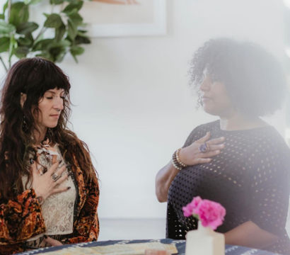 Two women in a cozy indoor spiritual session, hands over their hearts during a calm tarot reading with tarot cards, crystals and a patterned tablecloth, potted plant and framed art in the background.