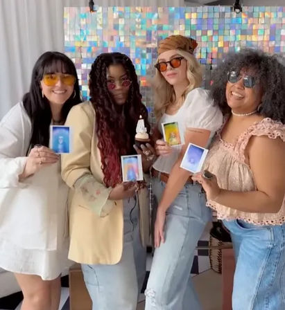Four people strike playful poses at an indoor photo booth with a holographic sequin backdrop, each holding a Polaroid-style instant photo while one holds a cupcake, wearing casual outfits and sunglasses.