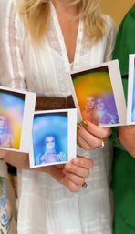 Close-up of hands holding instant Polaroid portraits with vibrant rainbow aura overlays, person in a white dress in the indoor background.