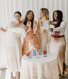 Four smiling women in pastel boho dresses holding Polaroid instant photos at a joyful indoor gathering, standing behind a lace-covered table with candles, flowers and rosé drinks.