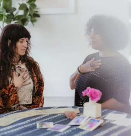 Two people with hands over their hearts in a bright spiritual healing session, tarot cards and crystals spread on a striped tablecloth with a pink flower