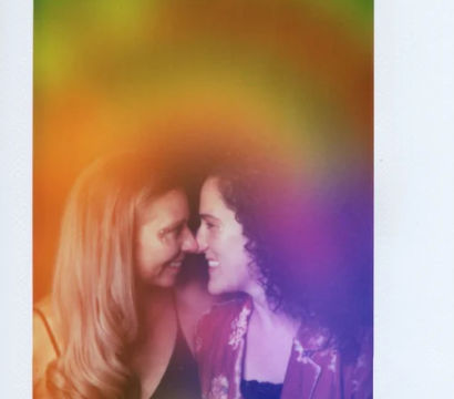 Instant-film portrait of two smiling women leaning forehead-to-forehead in an intimate pose, set against a vivid rainbow-colored light wash and white instant-film border.