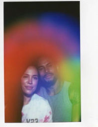 Portrait of two people posing closely, bathed in a dreamy rainbow haze with red, purple and green tones