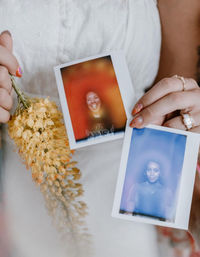 Close-up of hands with colorful nail art and rings holding two Polaroid instant portraits—one warm red-toned smiling face and one cool blue-toned portrait—beside a dried yellow flower bouquet against a white eyelet dress.