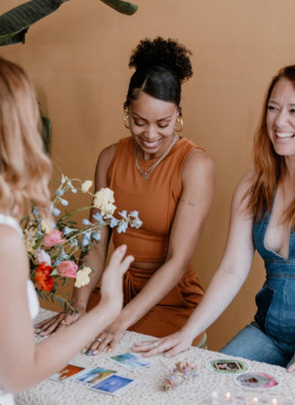 Three women smiling during a cozy boho tarot card reading at an indoor table with flowers, crystals, and candles