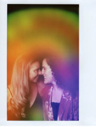 Polaroid-style portrait of a joyful same-sex couple nose-to-nose, smiling in a vibrant rainbow gradient light overlay — intimate, colorful romantic photo.