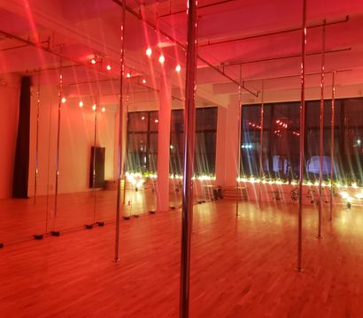 Pole dance studio loft with mirrored wall and multiple chrome poles, warm red ambient lighting, hardwood floors, string lights and potted plants along large industrial windows.