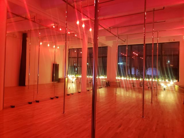 Pole dance studio loft with mirrored wall and multiple chrome poles, warm red ambient lighting, hardwood floors, string lights and potted plants along large industrial windows.
