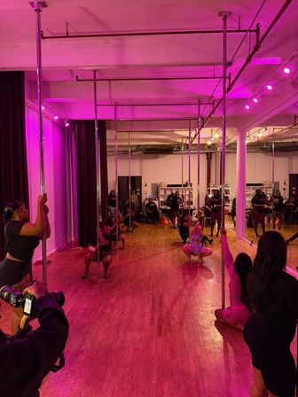 Energetic pole fitness class in a pink-lit studio — chrome poles, hardwood floor and mirrored wall with participants practicing moves while a photographer shoots.